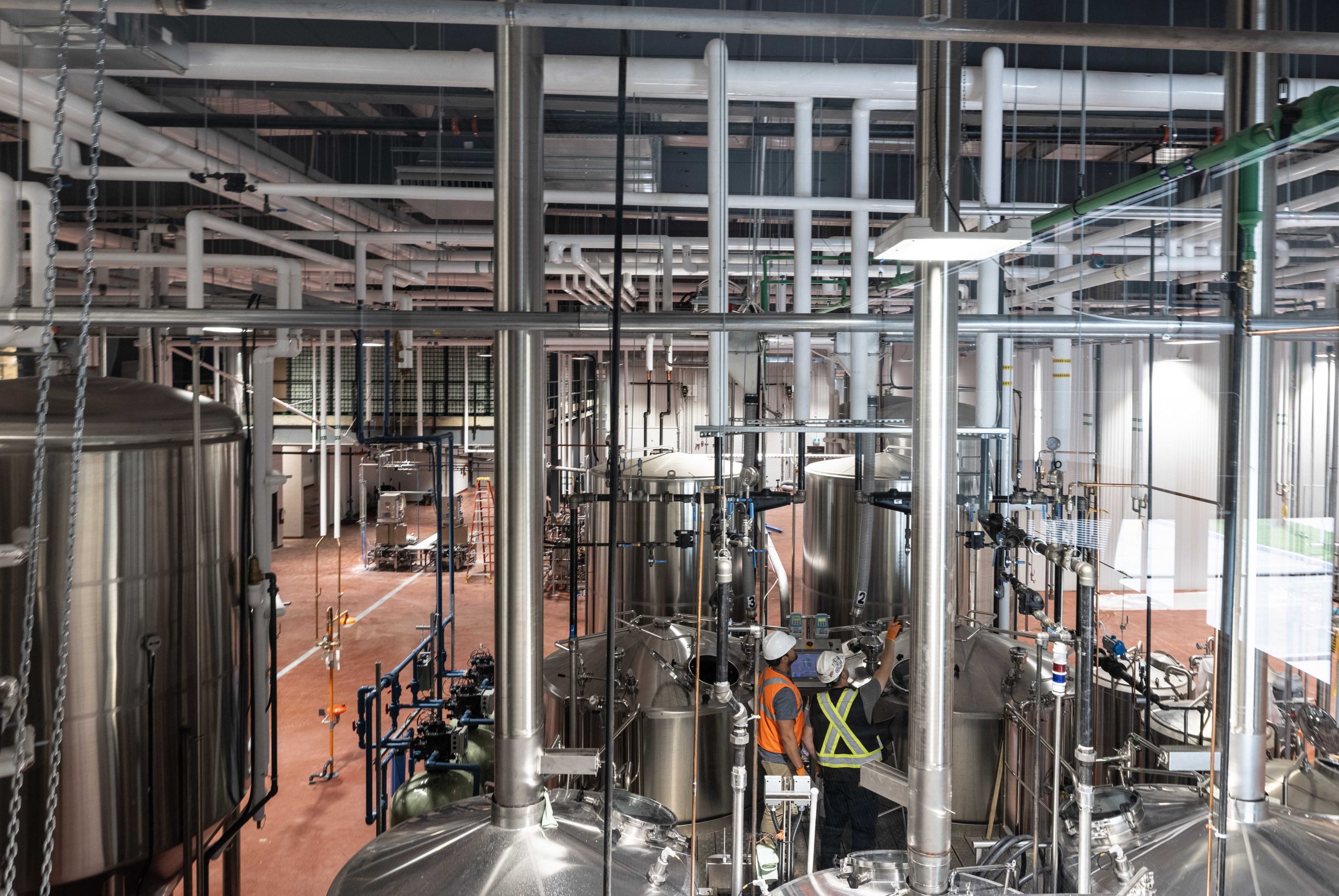 Brewery interior with large stainless steel tanks, pipes crisscrossing overhead. A worker in an orange vest inspects equipment, conveying industriousness.