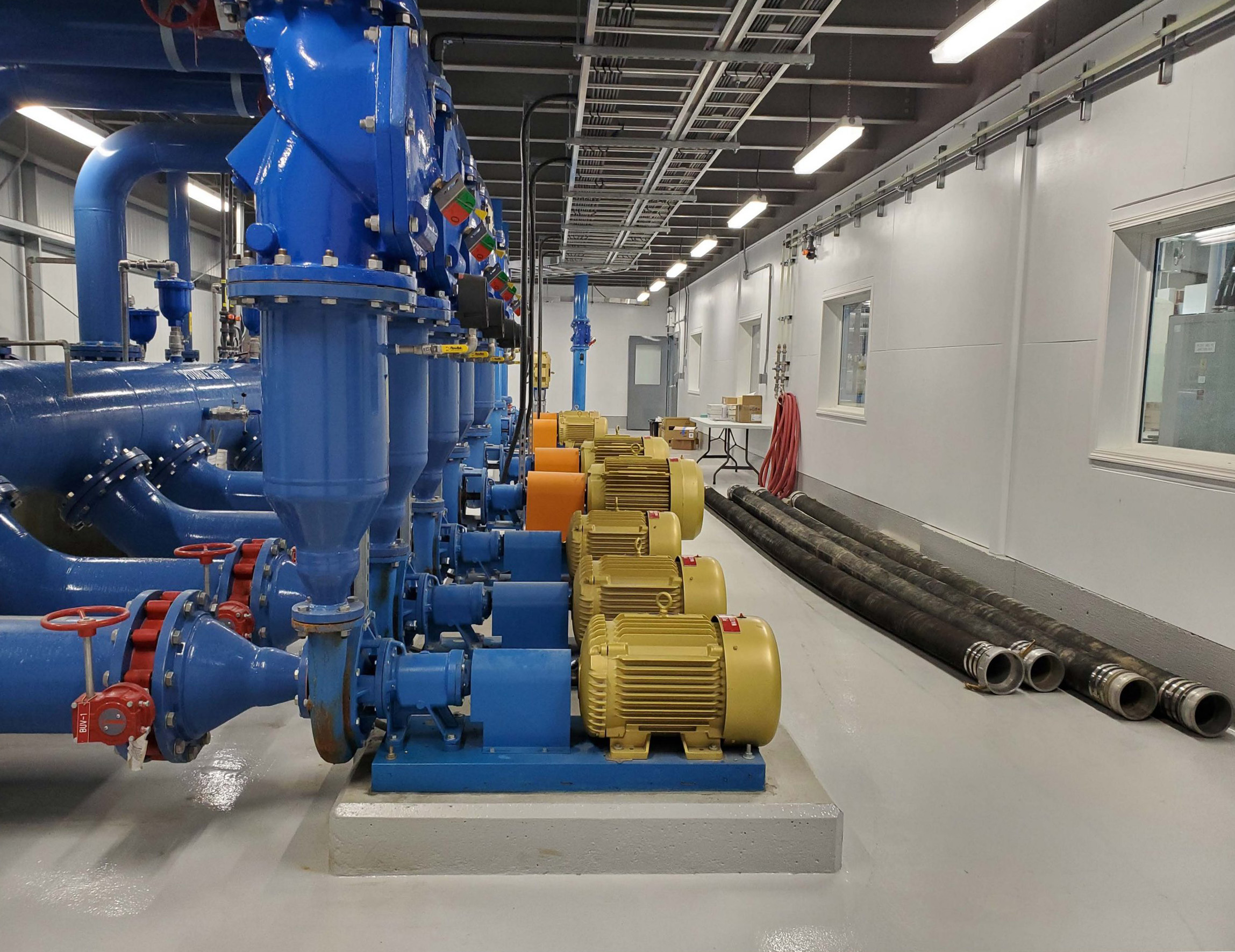 Industrial room with large blue pipes and yellow motors lined up on the left. Coiled hoses and black pipes rest along a clean white floor.