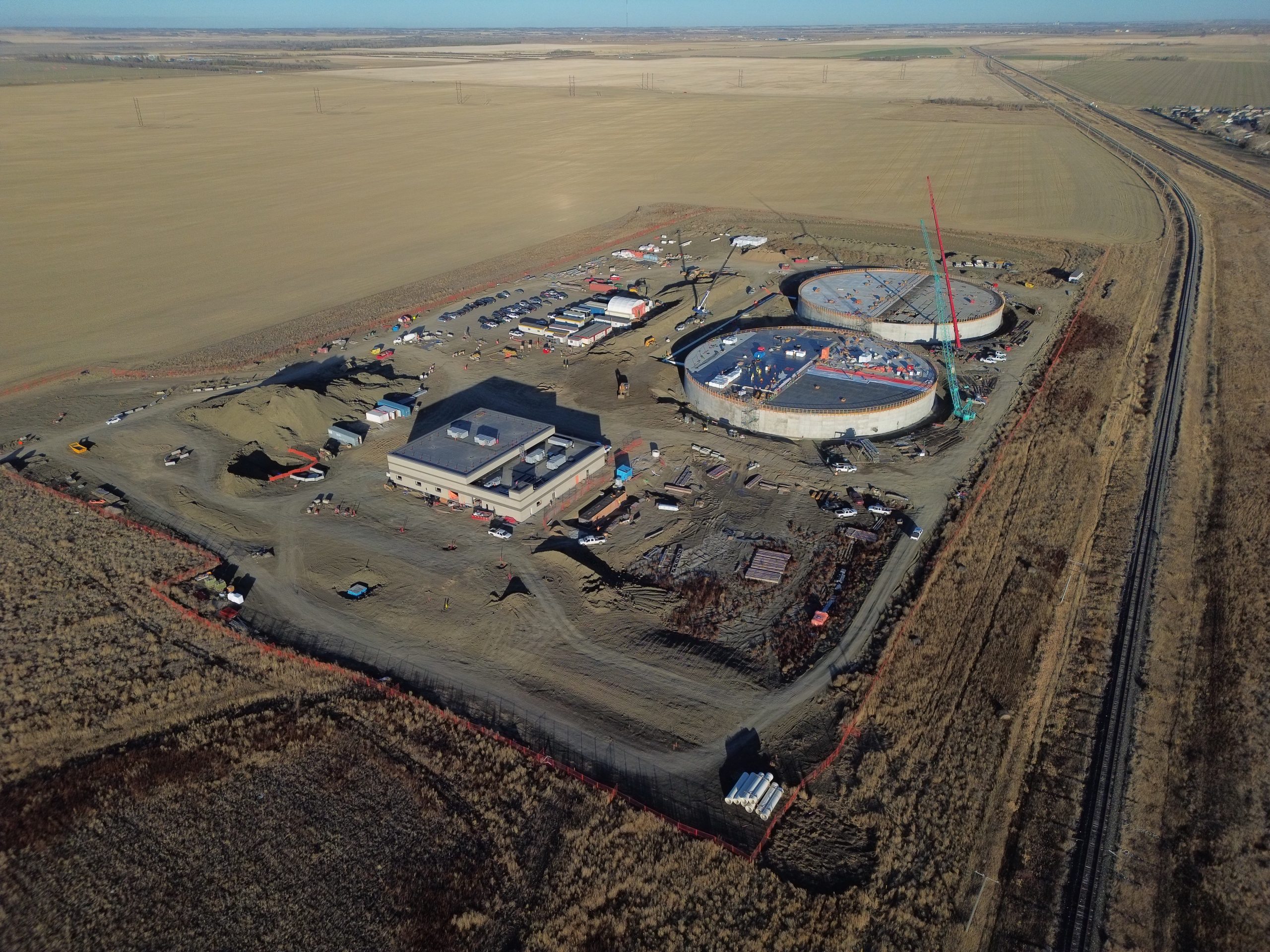Aerial view of a remote construction site with two large circular structures and several smaller buildings surrounded by barren land and dirt roads.