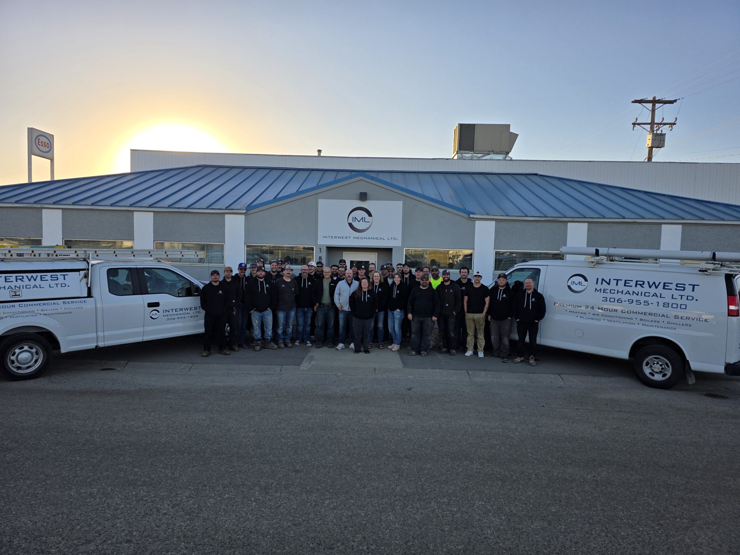 A group of people stand between two white vans with "Interwest Mechanical Ltd." logos, in front of a building at sunrise, conveying teamwork.